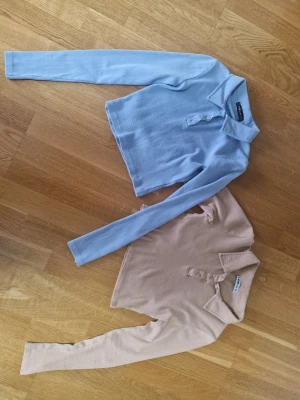 Två croppade långärmade polotoppar - Säljer två croppade långärmade polotoppar från Ginatricot. En är ljusblå och den andra beige, båda i ribbad bomull med krage och knappar framtill. Perfekta till jeans eller kjol för en chill look.