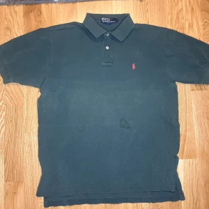 Svart piké från Ralph Lauren - Klassisk mörkgrön pikétröja från Ralph Lauren med två knappar vid kragen och den ikoniska röda broderade loggan på bröstet. Tillverkad i bomull med korta ärmar och normal passform. Perfekt till jeans eller shorts för en clean look.