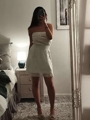 Vit axelbandslös kortklänning - Säljer en söt vit axelbandslös kortklänning med volang upptill och spetsdetaljer längst ner. Klänningen har en smockad midja som ger en fin passform och är perfekt för varma dagar.