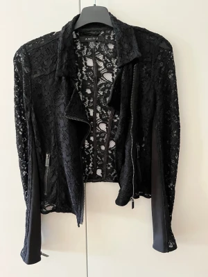 Svart spetsjacka från Amisu - Säljer en svart jacka från Amisu i genombruten spets med coola dragkedjedetaljer framtill och vid ärmsluten. Jackan har lång ärm, klassisk krage och en riktigt snygg, feminin look. Perfekt för dig som vill sticka ut lite extra. Liten i storlek så mer en 32/XXS