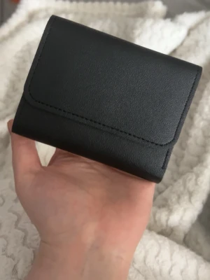 Svart plånbok i skinn - En liten svart plånbok i skinn material. Kommer nog gå ner i priset sen men säljer nu 21kr då de kommer med fri frakt just nu o vet inte hur mkt frakten är annars ju. De är ett litet hål i båtten i de stora facket som går att se på sista bilden men de va så jag köpte den och den är nog där för att kunna vika plånboken bättre .