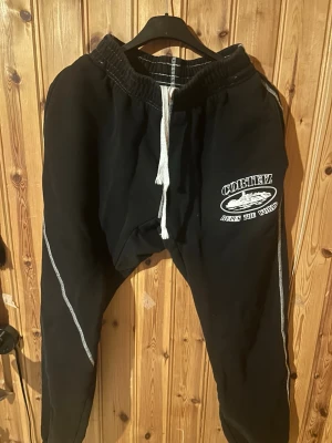 Svarta Cortez mjukisbyxor med snörning - Svarta mjukisbyxor från Cortez med vit snörning i midjan och tryck på ena benet. Byxorna har kontrastsömmar längs benen och baktill, samt en bakficka. Perfekta för chill dagar eller träning.