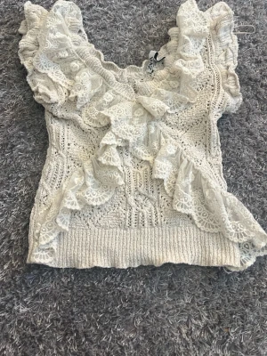 Vit spetsvolang topp från Zara - Supersöt vit topp från Zara i stickad bomull med breda spetsvolanger framtill och på axlarna. Toppen har en feminin och romantisk look med detaljerad spets och ribbad nederkant. Perfekt för dig som gillar en unik och söt stil. 