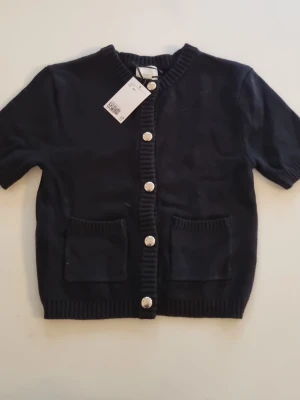 Svart kortärmad kofta från H&M - Kortärmad svart kofta från H&M. Koftan har ribbade kanter, två fickor framtill och stora silvriga knappar längs hela framsidan. 