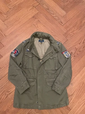 Fildjacka  - Säljer en grön parkajacka från Polo Ralph Lauren med coola patchar på ärmarna och flera fickor framtill. Jackan har dragkedja och knappar, samt klassisk krage. Perfekt för dig. 