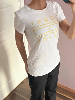 Vit Tommy Jeans t-shirt med gul print - Säljer en vit t-shirt från Tommy Jeans med gul grafisk logga framtill. Klassisk rund halsringning och korta ärmar. Skön passform och tillverkad i mjuk bomull. Perfekt till jeans eller shorts för en clean och enkel look.