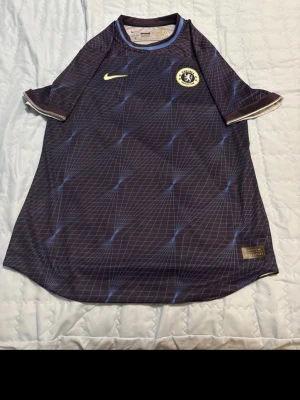 Chelsea Nike matchtröja blå XXL - Chelsea FC matchtröja från Nike i mörkblått med ljusblå rutmönster och guldfärgade detaljer. Tillverkad i lätt och ventilerande Dri-Fit polyester. Ikoniskt Chelsea-märke på bröstet och Nike Swoosh. Perfekt för dig som älskar fotboll och vill ha en snygg supportertröja.