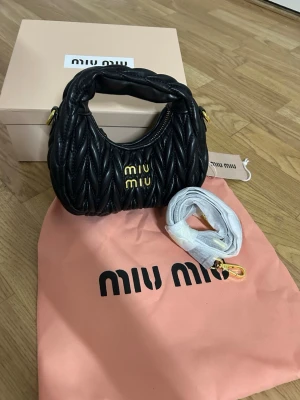 Svart quiltad handväska från Miu Miu - Supersnygg svart handväska från Miu Miu i quiltad skinn med guldfärgade detaljer och logga framtill. Väskan har en rundad form, dragkedja upptill och medföljande axelrem. Perfekt accessoar för att lyfta din outfit.