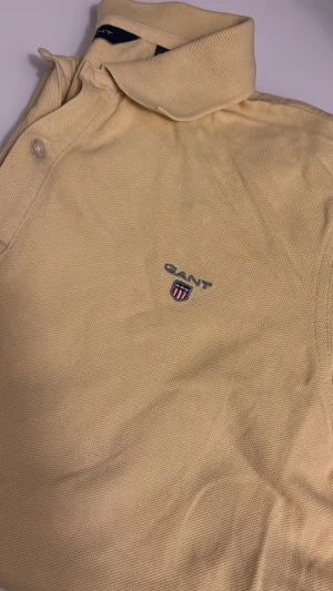 Gul pikétröja från Gant - Klassisk gul pikétröja från Gant med broderad logga på bröstet. Tröjan har krage, knappar vid halsen och är tillverkad i ett strukturerat bomullstyg.