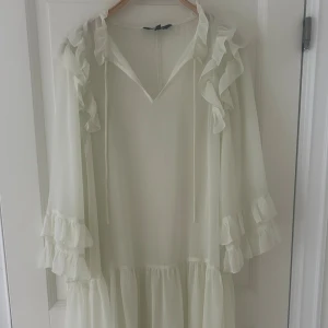 Vit volangklänning från KappAhl - Aldrig använd pga för liten!!Supersöt vit kortklänning från KappAhl med volanger längs ärmar och axlar. Klänningen har v-ringning med knytband och transparenta, flödande ärmar med flera lager volanger. Tillhörande underklänning i samma färg ingår. 