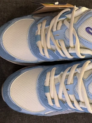 Vita och ljusblå Asics sneakers - Säljer ett par fräscha Asics sneakers i vitt och ljusblått. Skorna har mesh- och syntetpaneler för bra andningsförmåga och komfort. Klassisk snörning och en rejäl, mönstrad sula för bra grepp. Perfekta för dig som gillar sportig stil. Ja köpte dem och dem va små för mig 