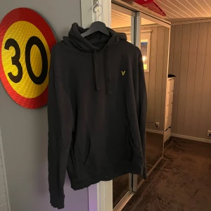 Grå hoodie från Lyle & Scott - Grå hoodie från Lyle & Scott med klassisk gul logga på bröstet. Tröjan har huva med snörning och en stor magficka framtill. Mjuk bomullskänsla och långärmad modell, perfekt för chill dagar. Snygg och enkel design som funkar till det mesta. Det är en XL men känns som en L.