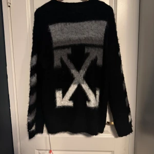 Off-White svart fluffig tröja - Svart fluffig stickad tröja från  med ikoniska vita och grå pilar på ryggen och diagonala ränder på ärmarna. Oversized passform och långärmad modell, perfekt för dig som gillar streetwear med statement. 