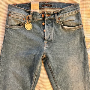 Blå jeans från Nudie Jeans, straight fit - Snygga blå jeans från Nudie Jeans, helt nya! Med klassisk femficksdesign och orange sömmar på bakfickorna. Modellen har raka ben och knäppning med knappar framtill. Jeansen är tillverkade i 100% bomull. Modell -  Grim Tim i storleken w31 L32.  Nypris 1600kr! säljer för ändast 799kr! W31/L32