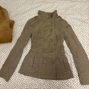 Beige jacka - Jackan har dubbelknäppning med hög krage och figurnära passform. 