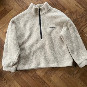 Beige fleece half zip från Pulling - Mysig beige fleece-tröja från Pulling med half zip och hög krage. Tröjan har dragsko nertill och är tillverkad i fluffigt syntetmaterial. Perfekt för kyliga dagar och enkel att styla med jeans eller mjukisbyxor.