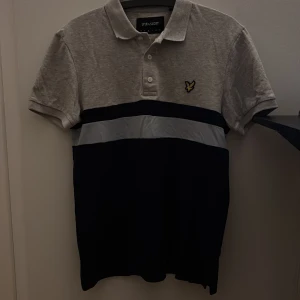 Grå och marinblå piké från Lyle & Scott - Snygg pikétröja från Lyle & Scott i grått och blått. Klassisk krage, korta ärmar och knappar framtill. Ikonisk gul logotyp på vänster bröst. Perfekt för dig som gillar stilren design med sportig touch.