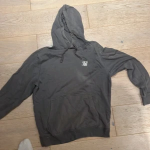 Grå hoodie från SikSilk med broderad logga - Mörkgrå hoodie från SikSilk med vit broderad logga på bröstet. Tröjan har huva med snörning, känguruficka och är tillverkad i mjuk bomullsmix. Perfekt för chill dagar och streetstyle.