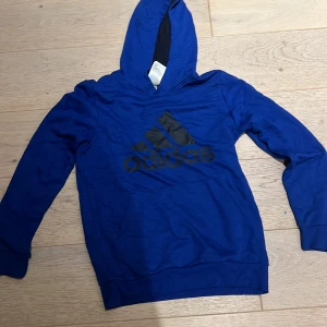 Blå Adidas hoodie med svart logga - Snygg blå hoodie från Adidas med stor svart logga på bröstet. Tröjan har huva, magficka och långa ärmar. Perfekt för dig som gillar sportig stil och vill ha något bekvämt och stilrent.