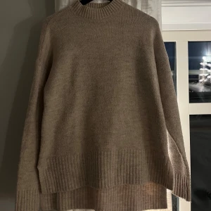 Beige stickad tröja med slits - Mysig beige stickad tröja med ribbad hög hals och breda ribbade muddar. Tröjan har en längre passform med slitsar i sidorna, perfekt för lager på lager. Långärmad och loose fit för en chill vibe. Tröjan är i storlek XS men sitter som en S.