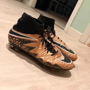 Nike Hypervenom fotbollsskor brons/svart - Säljer ett par Nike Hypervenom fotbollsskor i brons och svart med coolt geometriskt mönster och högt strumpliknande skaft. Skorna har snörning, platt sula med dobbar och är tillverkade i syntetmaterial. 