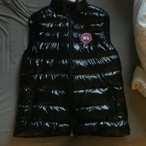 Svart dunväst från Canada Goose XL - Säljer en svart, glansig dunväst från Canada Goose i storlek XL. Västen har ståkrage, två sidofickor med dragkedja och det klassiska Canada Goose-märket på bröstet. Perfekt för lager-på-lager och har en quiltad design som ger extra värme.