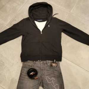 Svart hoodie från Polo Ralph Lauren - Svart hoodie från Polo Ralph Lauren i storlek L. Tröjan har huva med snörning, dragkedja framtill och två fickor. Klassisk vit broderad logga på bröstet. Mjuk bomullskänsla och stilren design som passar perfekt till jeans. Den har däremot små slitningar på slutet av armen 