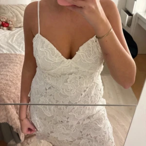 Vit spetsklänning med smala axelband - Supersnygg vit kort klänning med blommig spets och smala axelband. V-ringad framtill och figurnära passform. Spetsen har tydliga blommönster och klänningen har en fin kant nedtill. Perfekt som studentklänning exempelvis.