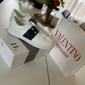 Valentino Garavani vita sneakers - Snygga vita sneakers från Valentino Garavani med bred marinblå detalj över sidan och tungan. Skorna har rund tå, platt sula med mönstrad undersida och klassisk snörning. Tillverkade i skinn med guldfärgad logga på insidan. Perfekt för dig som gillar stilrena designer. Stl 41 jätte fint skick. Påse och box medföljer. 