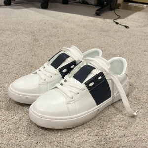 Valentino Garavani vita sneakers - Snygga vita Valentino Garavani sneakers med bred marinblå detalj över sidan och klassiska nitar på hälen. Skorna har rund tå, vita snören och är tillverkade i skinn för en lyxig känsla. Perfekta för dig som vill sticka ut med stilrena och exklusiva sneakers.