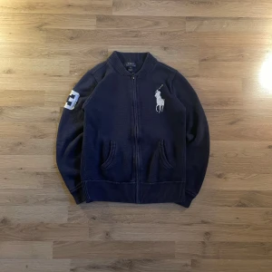 Mörkblå stickad cardigan jacka Polo - Snygg mörkblå stickad cardigan jacka från Polo Ralph Lauren med dragkedja framtill, ribbade muddar och två fickor. Stor vit broderad polospelare på bröstet och en vit siffra 3 på ena ärmen. Perfekt för dig som gillar klassisk stil med sportig touch. Passar på storlek S.  Bilder på måtten finns tillgängliga. Fråga gärna! Snabb leverans!
