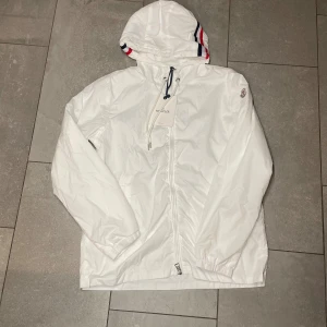 Vit vindjacka med huva - Snygg vit vindjacka med huva och coola röd-blå-vita ränder på kanten. Jackan har dragkedja framtill, snörning vid halsen logga på ärmen. Perfekt för dig som gillar stilrena och sportiga vibes.