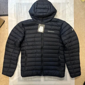 Svart Peak Performance dunjacka med huva - Svart dunjacka från Peak Performance med huva och diskret logga på bröstet och ärmen. Jackan har quiltad design, dragkedja framtill och två sidofickor med dragkedja. Perfekt för kalla dagar och har en clean, sportig look.