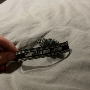 Tyg armband - Snyggt svart och silverfärgat armband med texten 'J'Adior' broderad. Armbandet har en stilren design med fransar i ena änden och är perfekt för dig som gillar exklusiva accessoarer med tydlig logga.