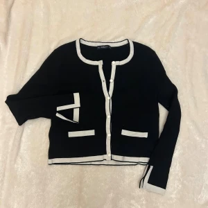 Svart kofta med vita detaljer från Zara - Stilren svart kofta från Zara med vita kontrastkanter och pärlknappar framtill. Koftan har två dekorativa fickor och långa ärmar med vita detaljer vid ärmslut. 