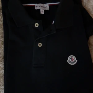 Svart Moncler pikétröja med logga - Svart pikétröja från Moncler i storlek S. Klassisk design med krage, två vita knappar och Moncler-logga broderad på bröstet. Tröjan är gjord i mjuk bomull och har en stilren look som passar perfekt till jeans eller shorts.