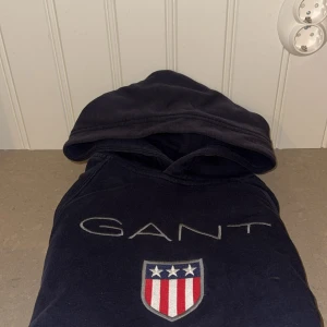 Mörkblå hoodie från GANT med logga - Säljer en mörkblå hoodie från GANT med broderad logga och sköld på bröstet. Tröjan har huva, magficka och långa ärmar. Klassisk och stilren design med vita och röda detaljer på loggan. 