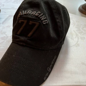 Sailracing keps - Svart keps från Sail Racing med broderad text och siffran 77 framtill. Kepsen har böjd skärm och justerbar passform. Tillverkad i bomull och har Sail Racing-logga broderad på sidan av skärmen. Perfekt för en sportig och avslappnad look.