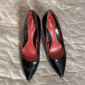 Svarta pumps med spetsig tå från Bershka - Snygga svarta pumps från Bershka med glansig finish och röd innersula. Skorna har spetsig tå och hög klack som ger en elegant siluett. Passar 38-39 Materialet är syntet med lackad yta och innersulan har dekorativ kant. Perfekt för dig som gillar att sticka ut.väldigt fina eleganta,Fynda!