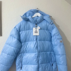 Ljusblå Moncler pufferjacka med huva - Säljer en ljusblå pufferjacka från Moncler med huva och dragkedja framtill. Jackan har två sidofickor med dragkedja, en ficka på ärmen med Moncler-logga och quiltad design. Insidan har en cool serietidningsdetalj. Perfekt för kalla dagar!