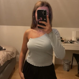 Ljusblå oneshoulder ribbad topp - Säljer en ljusblå ribbad topp med oneshoulder-design och lång ärm. Toppen har vågiga kanter vid ärmslut och nederkant, vilket ger en extra detalj. Materialet är mjukt och stretchigt, perfekt för dig som gillar en unik och stilren look. Aldrig använd