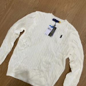 Vit kabelstickad tröja från Ralph Lauren - Kabelstickad vit tröja från Ralph Lauren med klassisk rund halsringning och broderad logga på bröstet. Tröjan har långa ärmar och ribbade muddar, perfekt för lager-på-lager. Snygg och tidlös design som funkar till allt.