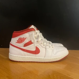 Nike Air Jordan 1 Mid vit/röd - Säljer ett par Nike Air Jordan 1 Mid i vitt och rött skinn. Klassisk siluett med röd swoosh, röda detaljer och Air Jordan-logga på sidan. Perforerad tå, snörning och röd yttersula. Perfekta för dig som gillar streetwear och sneakers med ikonisk design.