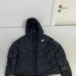 Svart jacka från Nike - Svart jacka från Nike med huva och vit logga på bröstet. Jackan har quiltad design, dragkedja framtill och långärmade ärmar. Perfekt för kalla dagar och har en clean, sportig look som funkar till streetwear. Priset kan diskuteras!