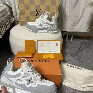 Louis Vuitton vita och grå sneakers - Säljer ett par exklusiva Louis Vuitton sneakers i vitt och ljusgrått med ikonisk blomdetalj på sidan. Skorna har snörning, platt sula och är tillverkade i en mix av skinn och textil. Kommer med originalkartong, dustbag och certifikat. Riktigt snygga och trendiga sneakers!