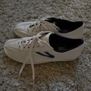 Vita sneakers från Tretorn - Säljer ett par klassiska vita sneakers från Tretorn med marinblå detaljer och snörning. Skorna är i canvasmaterial och har en platt sula samt rund tå. Perfekta för en clean och stilren look.