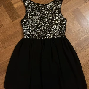 Svart paljettklänning med öppen rygg - Svart kortklänning i polyester och nylon med glittrig paljettöverdel i guld. Klänningen har ärmlös design, rund hals och en snygg öppen rygg med dragkedja bak. Perfekt för dig som vill sticka ut med en festlig look.