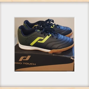 Oanvända Pro Touch fotbollsskor, strl 31 - Snygga svarta fotbollsskor från Pro Touch med limegröna detaljer och prickigt mönster på hälen. Skorna har snörning, rund tå och platt gummisula i brunt. Perfekta för inomhusspel och har en sportig look som sticker ut på planen.