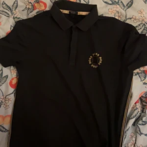 Svart kortärmad BOSS t-shirt med tryck - Svart t-shirt från BOSS med korta ärmar och klassisk krage. På bröstet finns ett gult BOSS-tryck i cirkel och längs sidan löper en snygg randig detalj i vitt och gult. Perfekt för dig som gillar stilrena märkesplagg med coola detaljer.