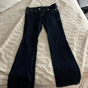 True Religion Becca Bootcut Jeans - Mörkblå jeans från True Religion, modell Becca Mid Rise Bootcut. Snygga kontrastsömmar och klassiska bakfickor med märkets ikoniska broderi. Jeansen har medelhög midja och bootcut-ben, tillverkade i jeansmaterial.